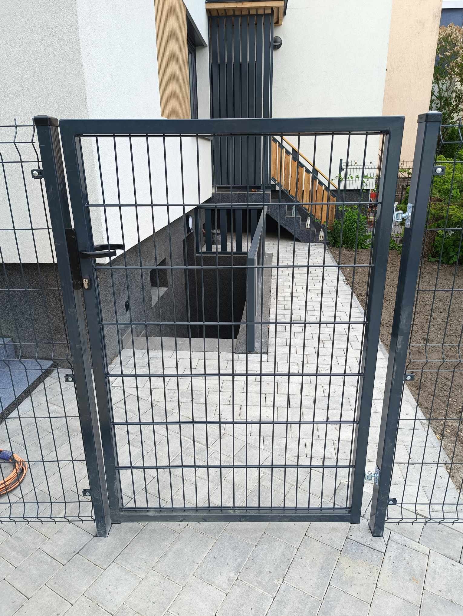 Nowoczesna brama z metalowej siatki w kolorze grafitowym, prowadząca do zejścia do piwnicy w nowym domu. Widoczna kostka brukowa i fragment ogrodzenia panelowego.
