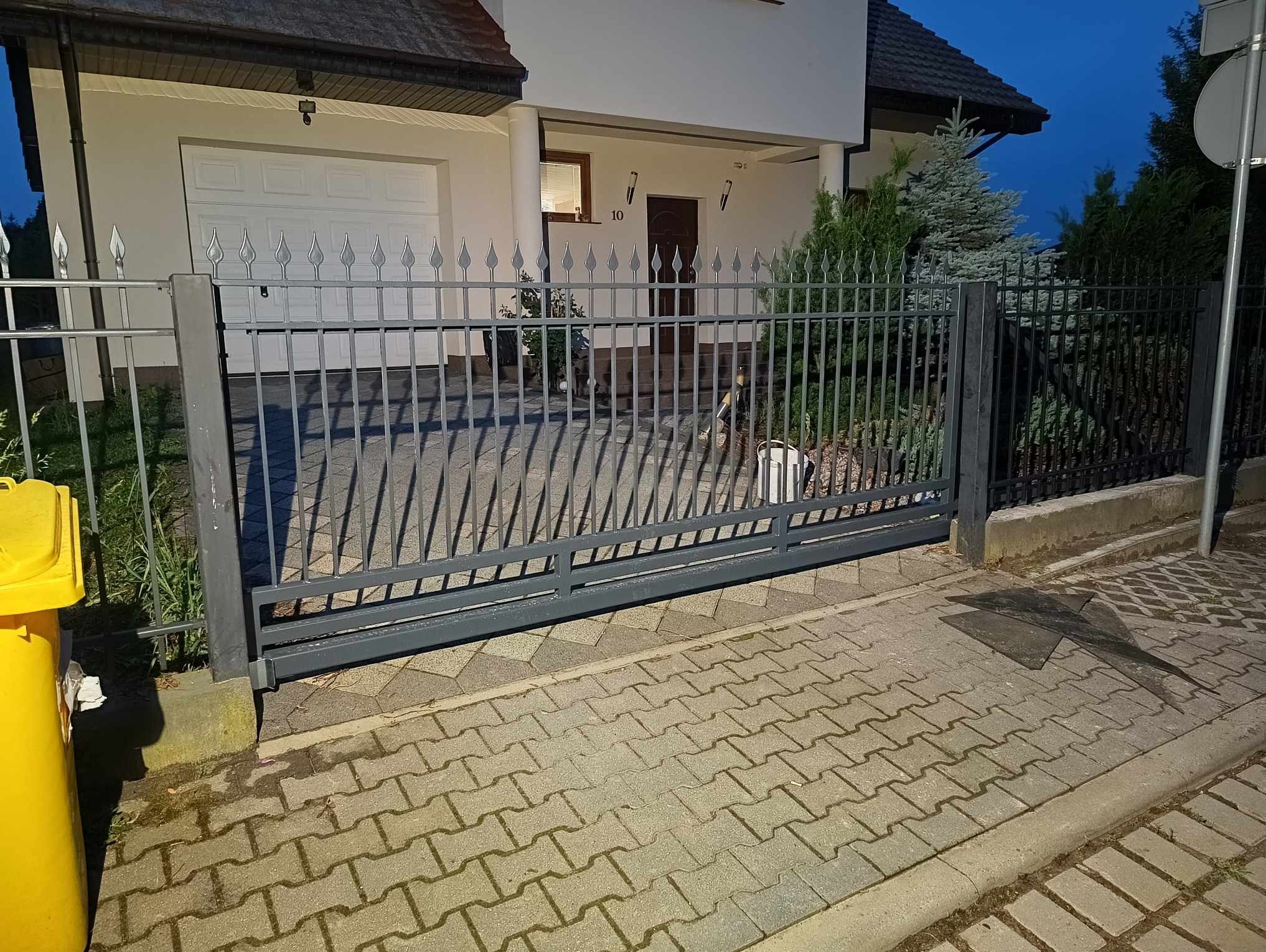 Przesuwna brama wjazdowa z metalowymi sztachetami w kolorze szarym, widok na dom z białymi ścianami i garażem, kostka brukowa na podjeździe, wieczorne oświetlenie.