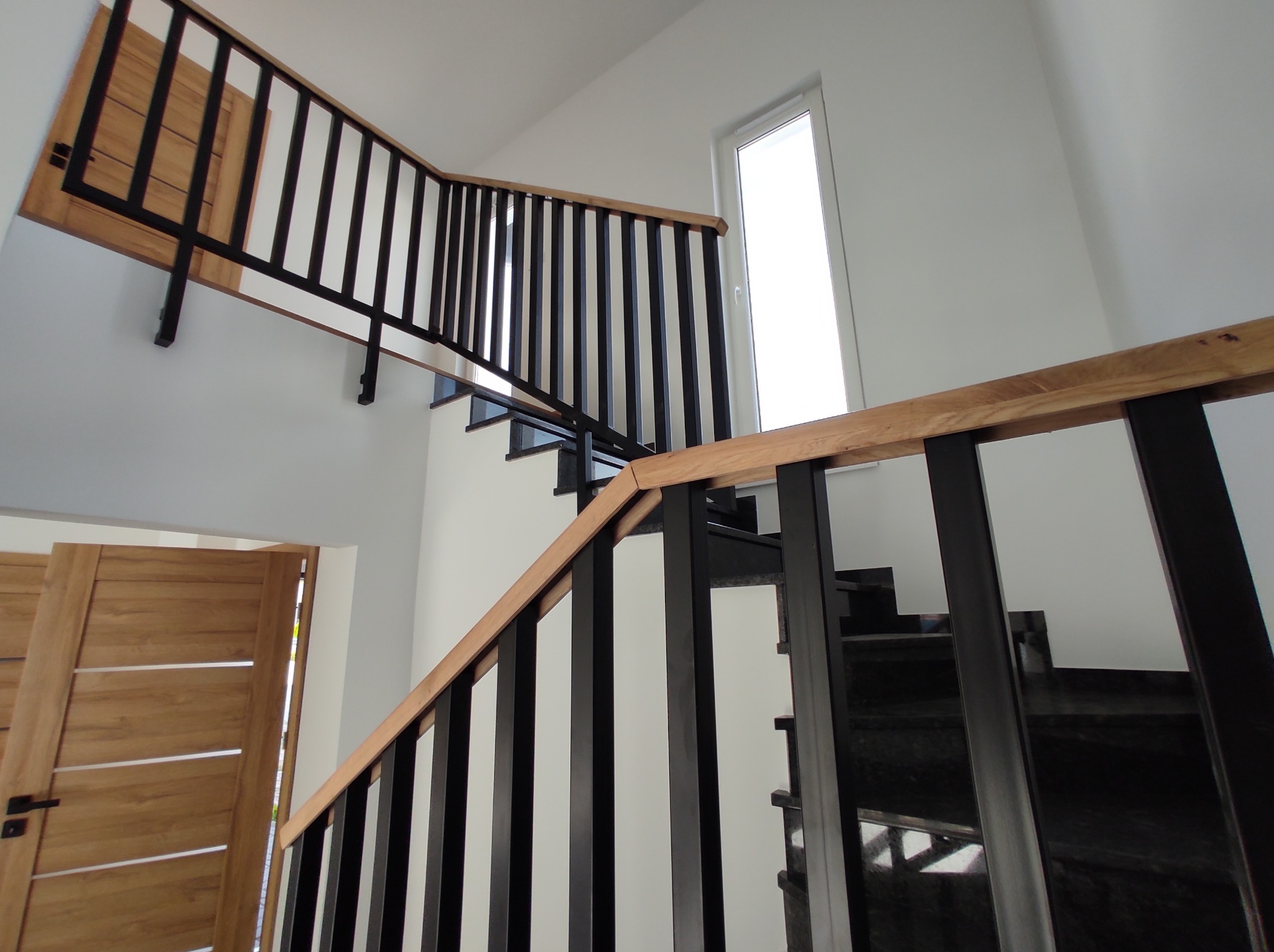 Nowoczesne schody z czarnymi metalowymi balustradami i drewnianymi poręczami, widok z dołu, z fragmentem drewnianych drzwi w tle.