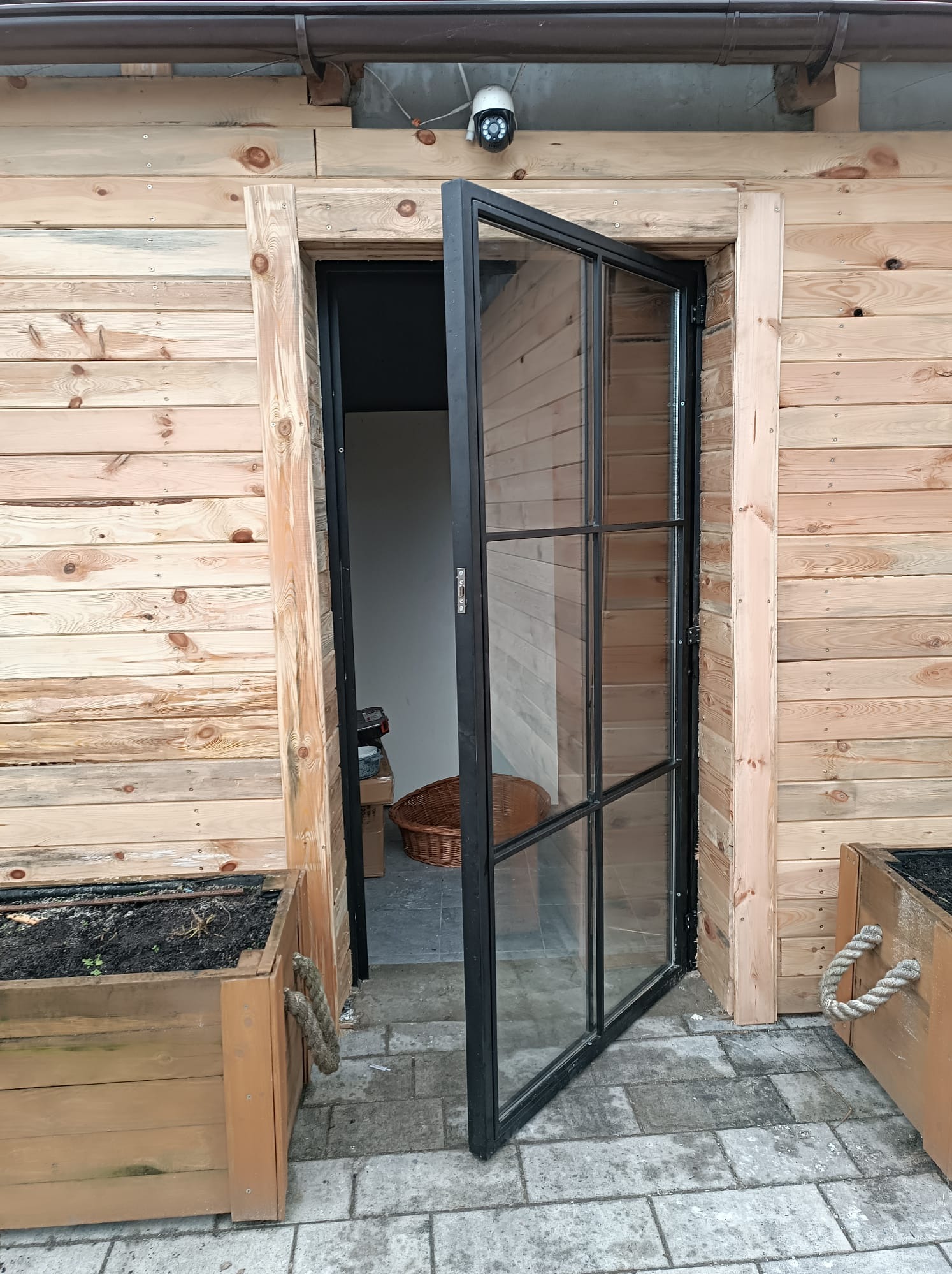 Otwarta, przeszklona, czarna metalowa rama drzwiowa w drewnianej ścianie budynku, widoczne donice z ziemią i sznurkowymi uchwytami po bokach.