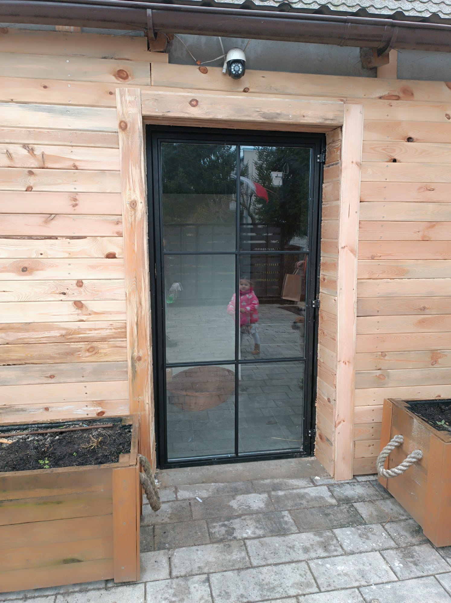 Nowoczesne drzwi z czarną metalową ramą i szklanymi panelami, wkomponowane w drewnianą elewację budynku z widocznymi skrzyniami na rośliny po bokach.