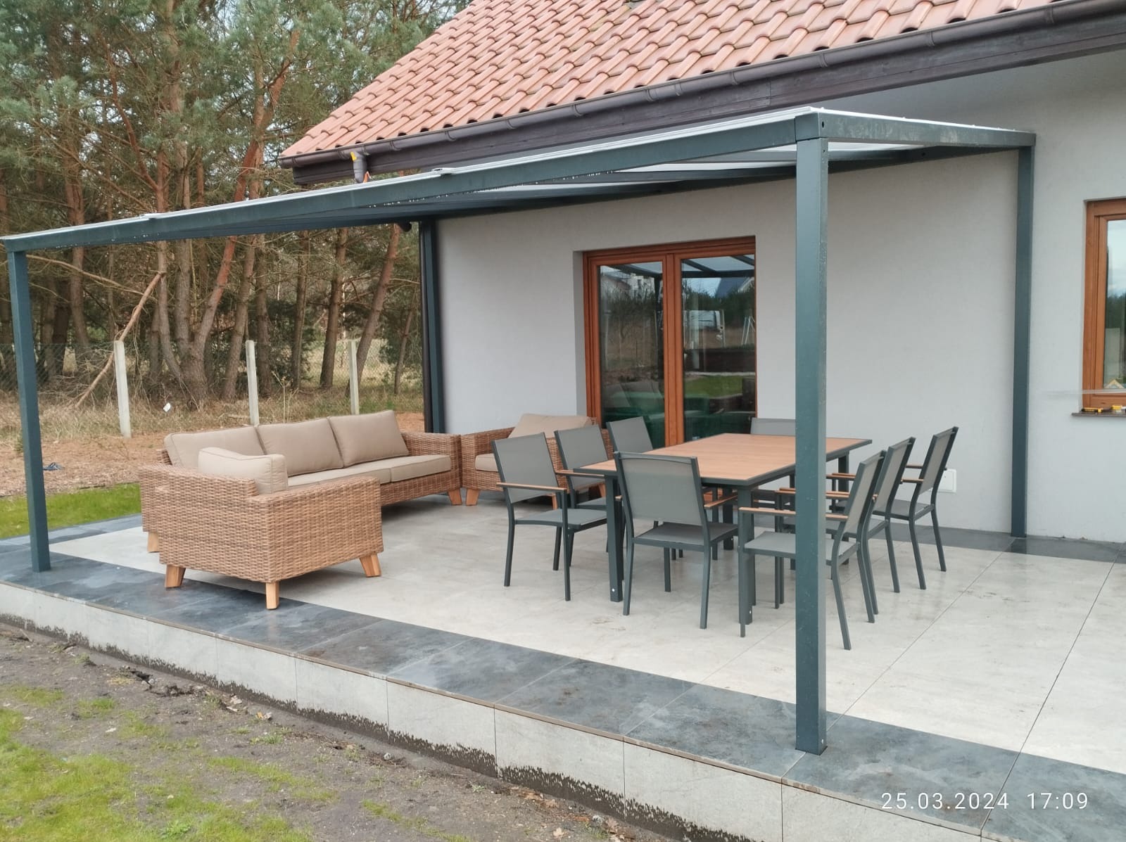 Zadaszony taras z metalową ramą pergoli, wyposażony w meble ogrodowe z rattanu i zestaw jadalniany z ciemnymi krzesłami, na tle jasnego domu z czerwoną dachówką i widokiem na drzewa.