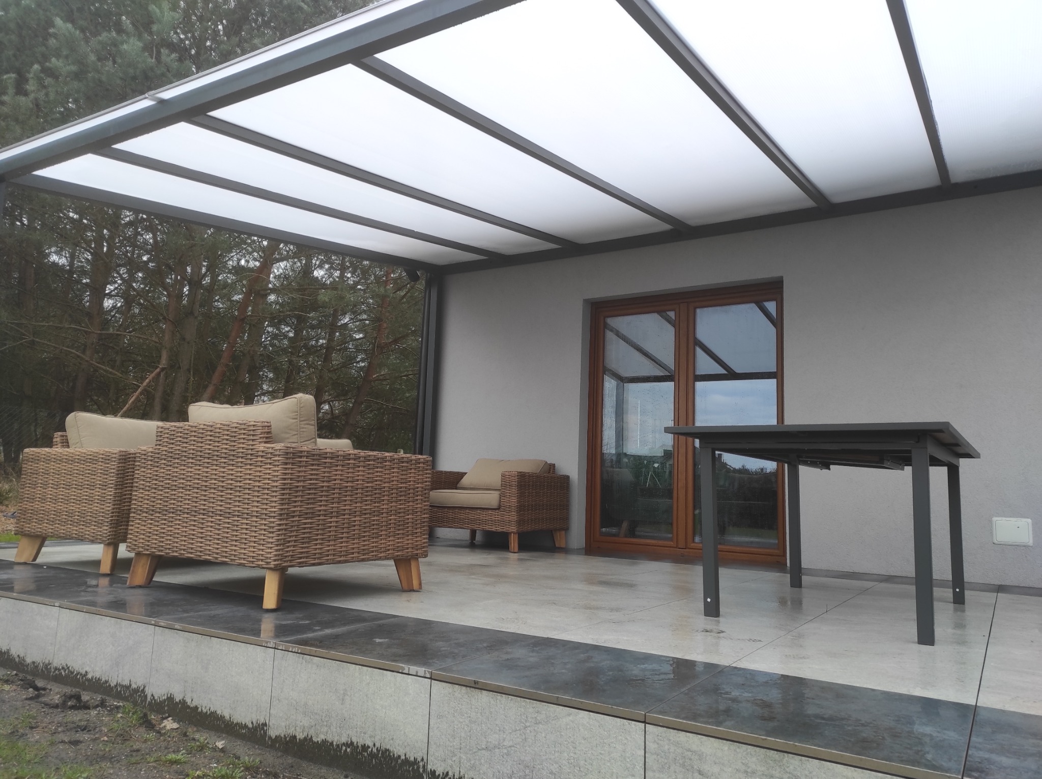 Zadaszony taras z meblami ogrodowymi z rattanu i metalowym stołem na płytkach, widok na ogród przez przeszklone drzwi i metalową konstrukcję zadaszenia.