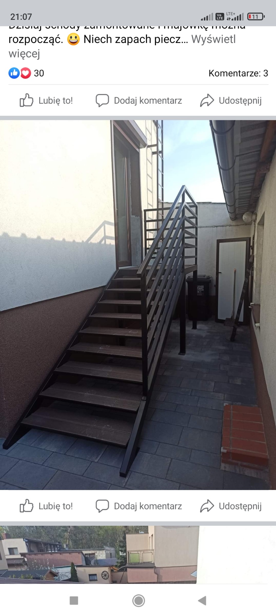 Zewnętrzne schody metalowe z drewnianymi stopniami prowadzące na balkon, widoczne z boku, z elementami zadaszenia i czarnym pojemnikiem na śmieci obok.