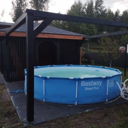 E-ALTANY - Niebieski basen Bestway Steel Pro pod czarną, drewnianą pergolą, obok ciemnej altany z pionowymi deskami, na szarym, gumowym podłożu.