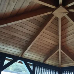 Wnętrze drewnianej altany z widoczną konstrukcją dachu z belek i desek, pomalowaną na brązowo, z czarnymi elementami ramy i widokiem na zewnątrz przez szczeliny w panelach.