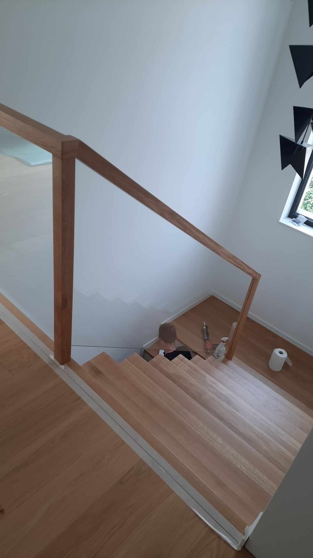 Jasne drewniane schody z przeszkloną balustradą i drewnianą poręczą, widok z góry na osobę schodzącą, obok leżą narzędzia i rolka papieru.