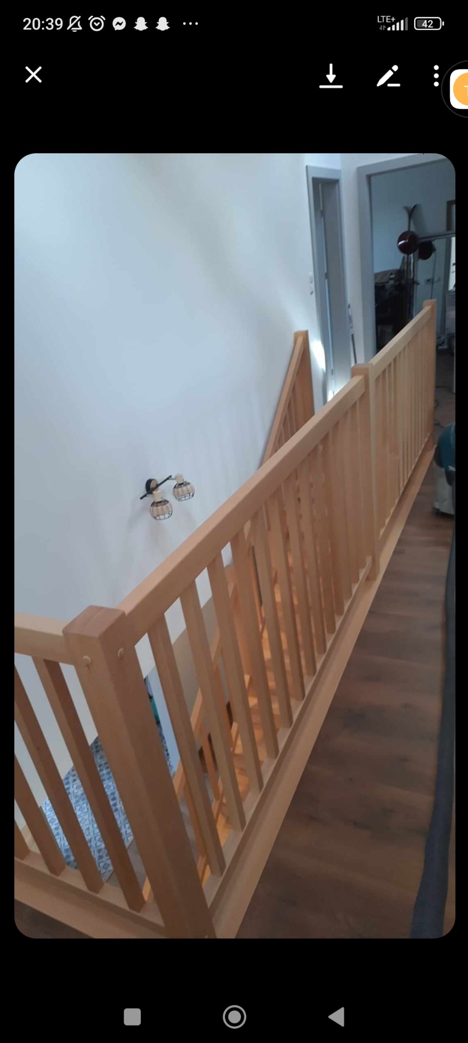 Jasne, drewniane schody z barierką w nowoczesnym wnętrzu, widok z góry na dół, z elementami oświetlenia na ścianie i widocznym fragmentem podłogi.