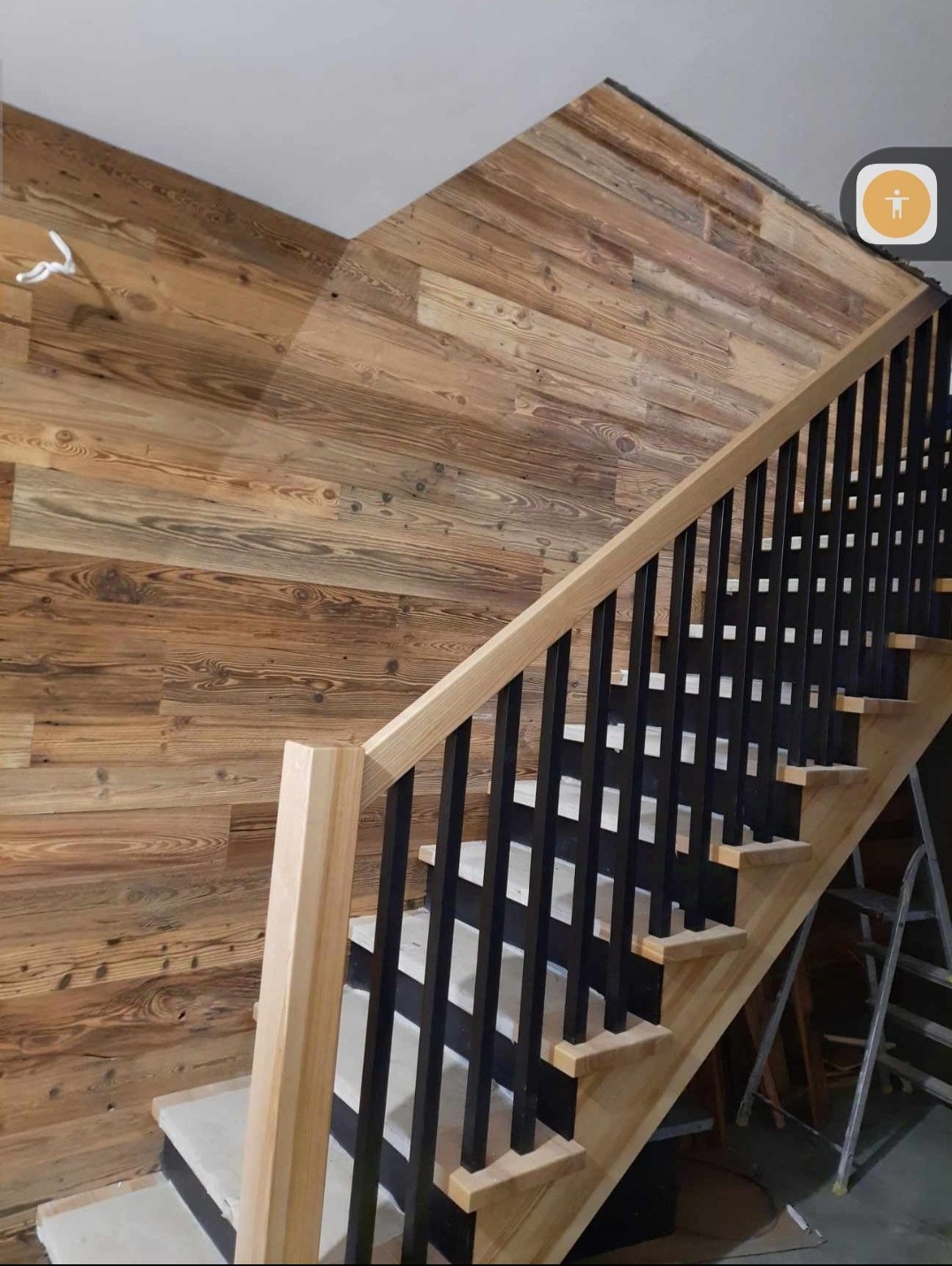 Drewniane schody z czarnymi balustradami, wykończone deską boazeryjną na ścianie, widok z dołu, drabina w tle.