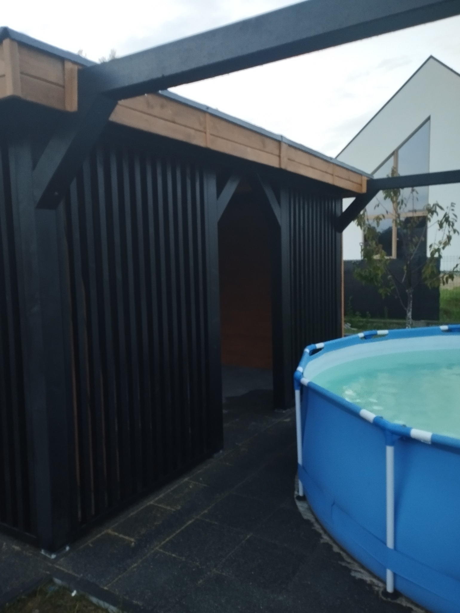 Czarna, drewniana konstrukcja z otwartym wejściem, prawdopodobnie szatnia basenowa, obok której stoi niebieski basen stelażowy z wodą, na tle domu z dużym oknem.