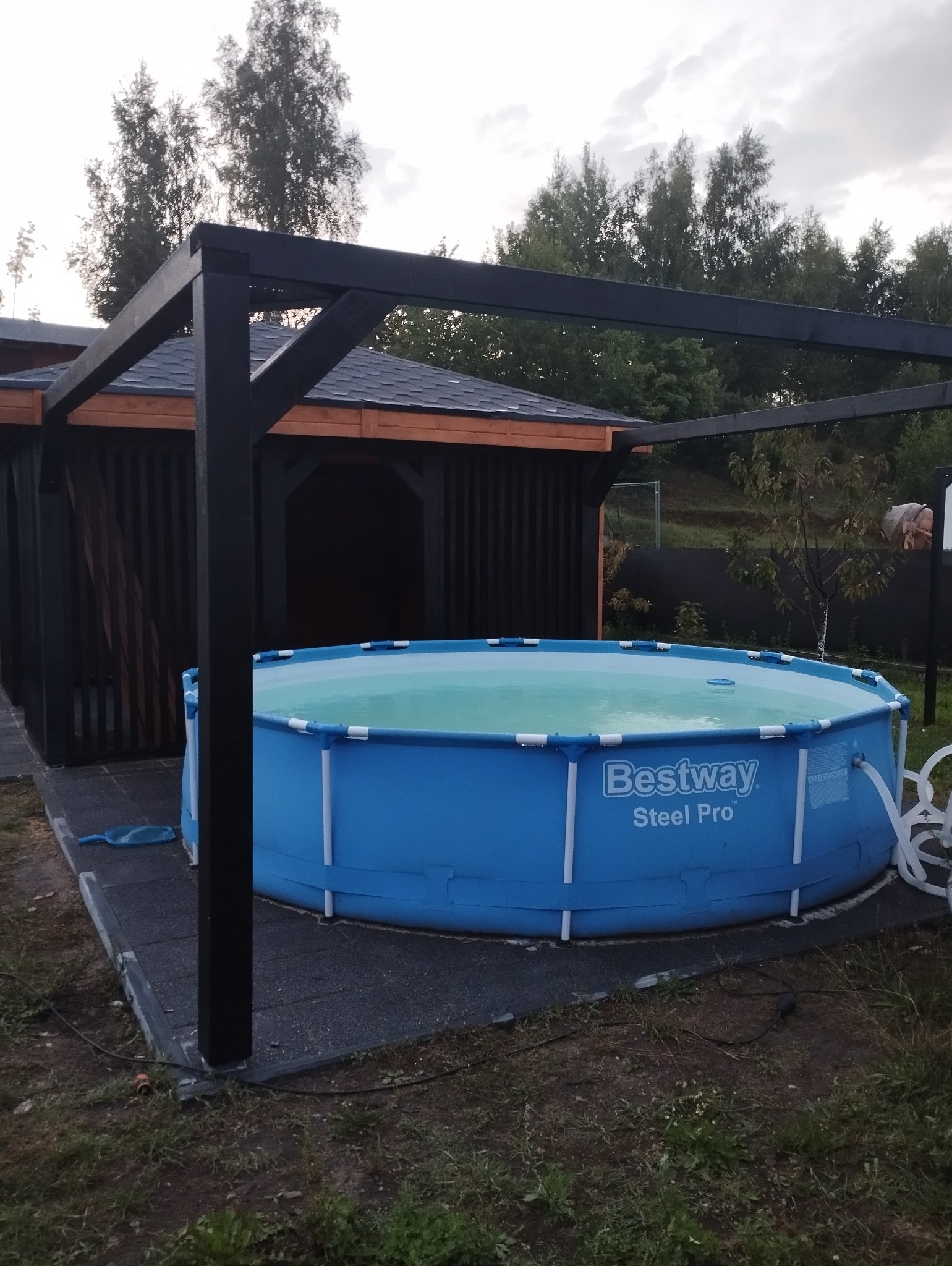 Niebieski basen Bestway Steel Pro pod czarną, drewnianą pergolą, obok ciemnej altany z pionowymi deskami, na szarym, gumowym podłożu.