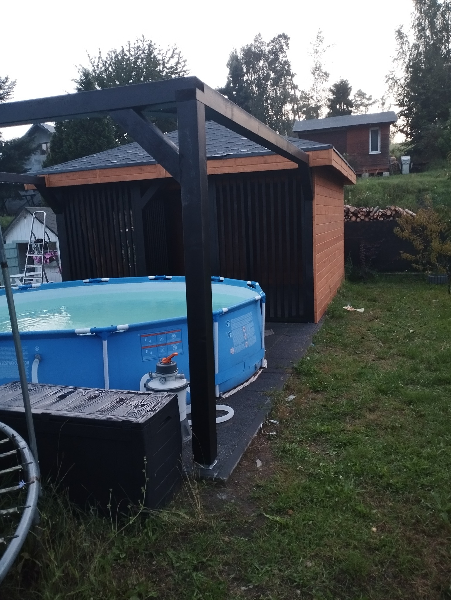 Zadaszenie nad basenem ogrodowym z czarną konstrukcją i drewnianą zabudową, widoczny basen z wodą, w tle domek letniskowy i stos drewna.