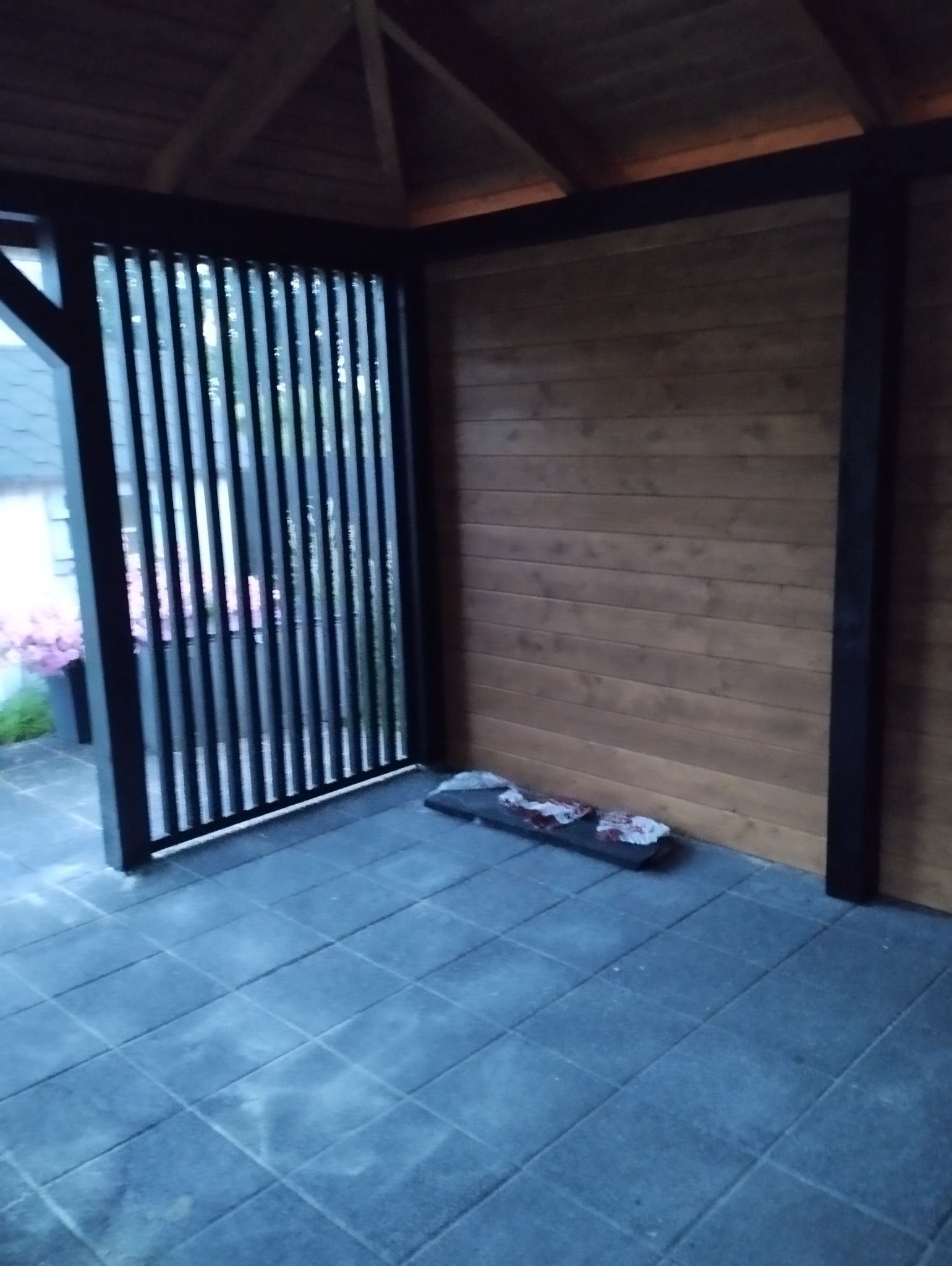 Zadaszona przestrzeń z drewnianą ścianą i ażurową, pionową przesłoną z czarnymi ramami, wyłożona szarą kostką brukową. W rogu leżą zabezpieczenia budowlane.