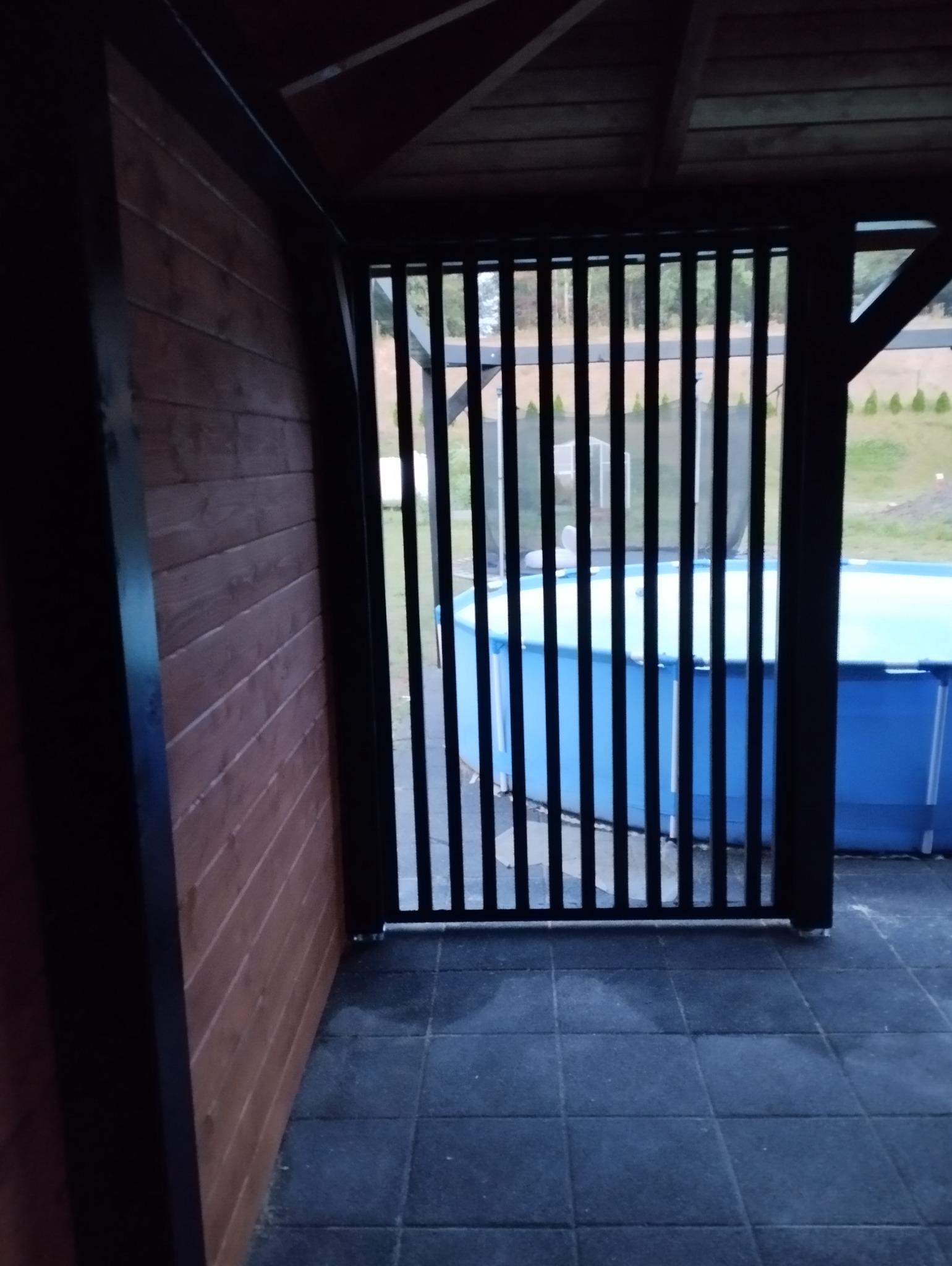 Zadaszona przestrzeń z drewnianą ścianą i czarną, pionową, drewnianą kratą, za którą widoczny jest basen ogrodowy.