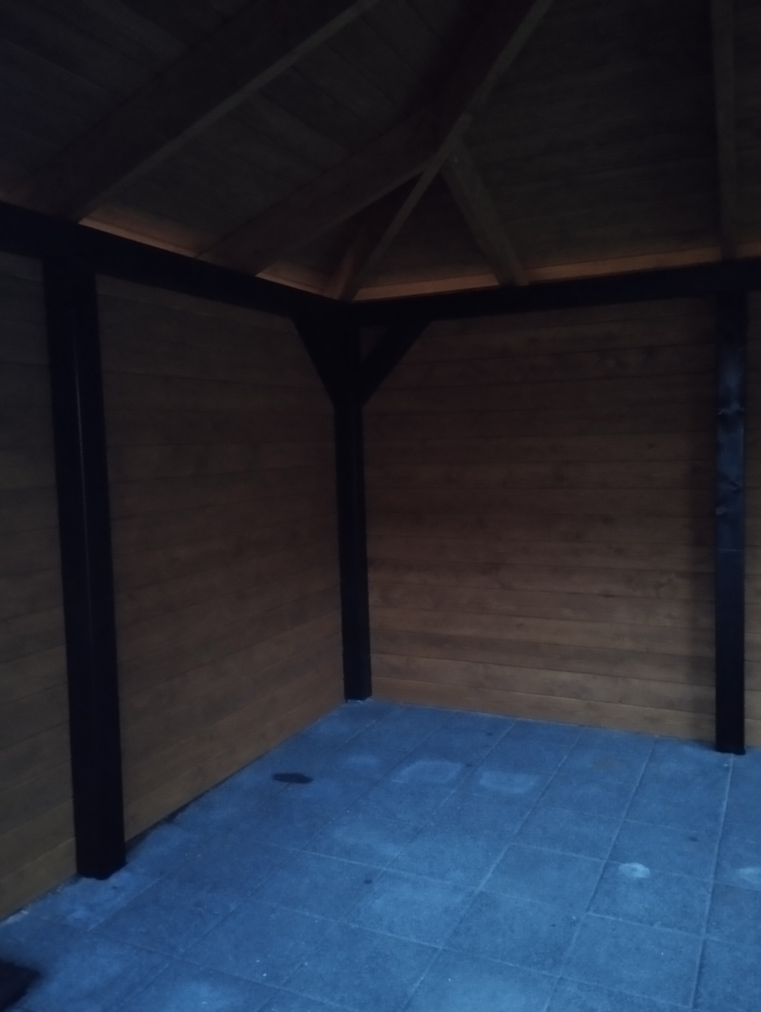 Wnętrze drewnianej altany z ciemnymi, malowanymi na czarno słupami i szarą kostką brukową na podłodze, widok z dołu na drewniany strop.