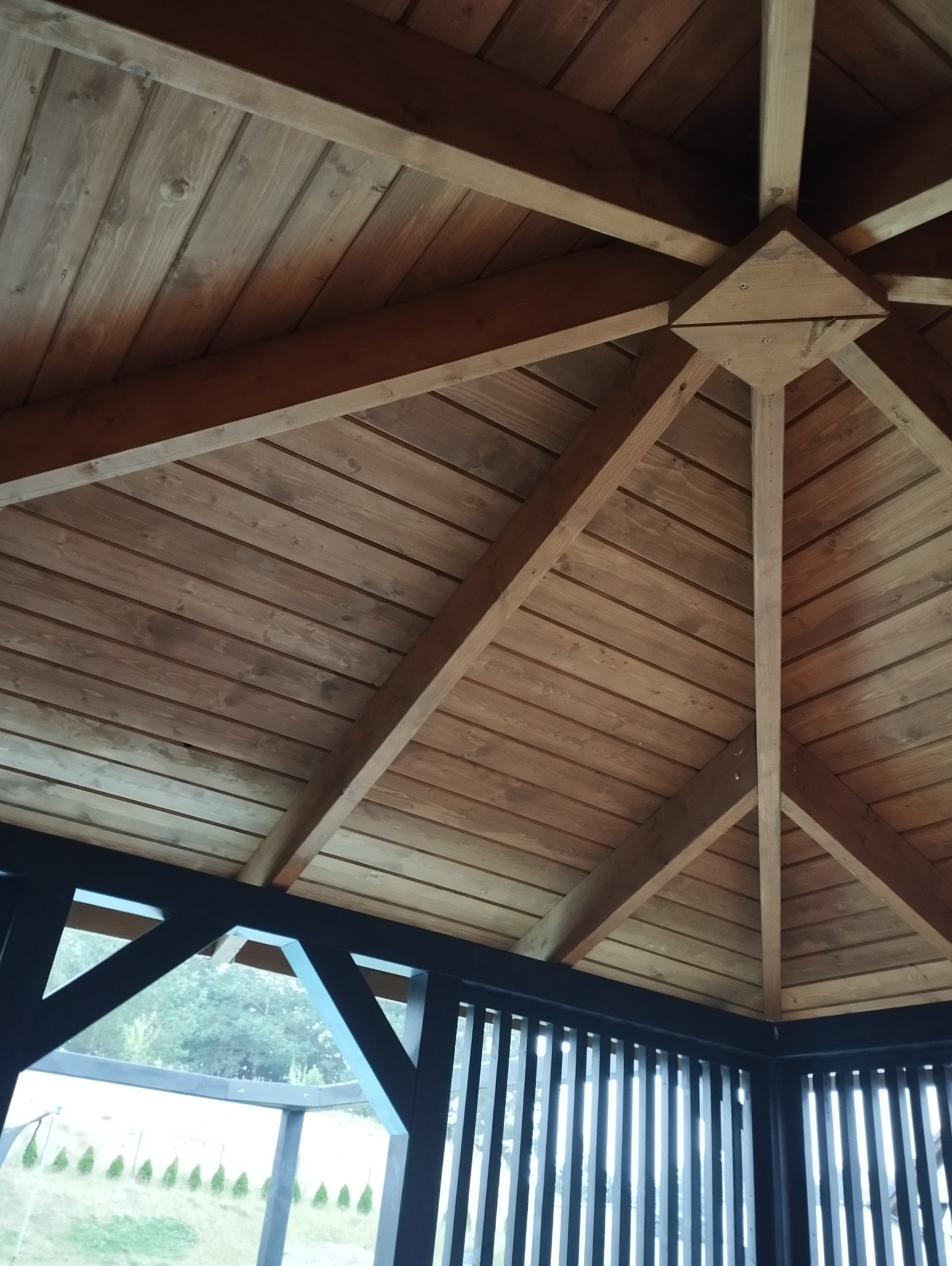 Wnętrze drewnianej altany z widoczną konstrukcją dachu z belek i desek, pomalowaną na brązowo, z czarnymi elementami ramy i widokiem na zewnątrz przez szczeliny w panelach.