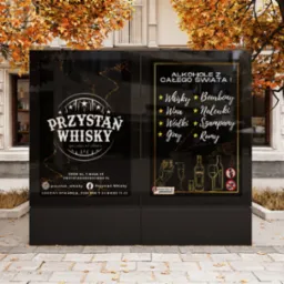 Billboard reklamowy z czarnym tłem prezentujący ofertę baru 'Przystań Whisky' z alkoholem z całego świata, umieszczony na chodniku w otoczeniu drzew w jesiennej scenerii.