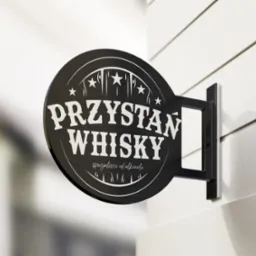 Okrągły, czarny szyld z napisem 'PRZYSTAŃ WHISKY specjaliści od alkoholu' zawieszony na białej ścianie z poziomymi panelami.
