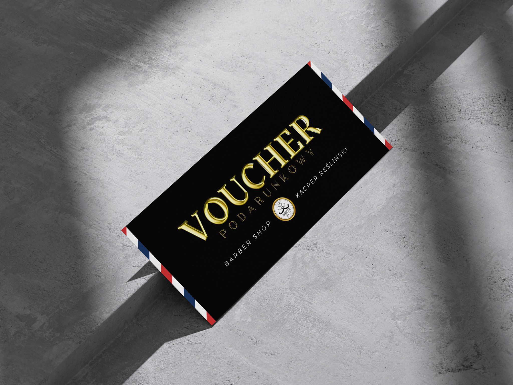 Czarny voucher prezentowy dla barber shopu z napisem 'Voucher Podarunkowy' w złotym kolorze, leżący na szarym, teksturowanym tle.
