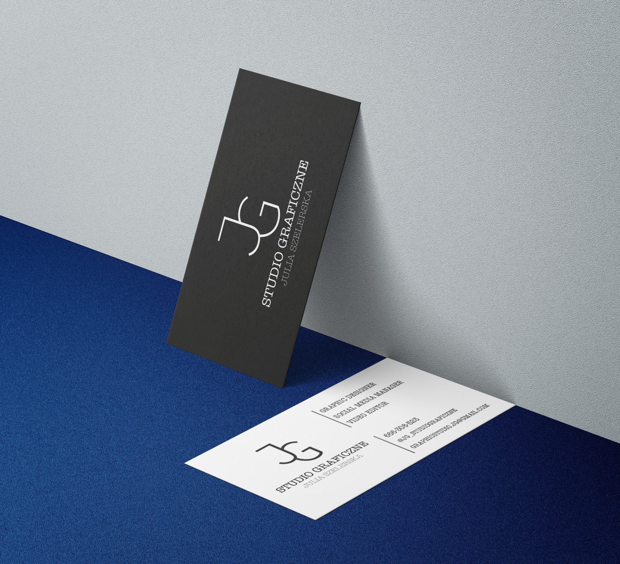 Dwie wizytówki na teksturowanym tle: jedna czarna pionowa oparta o ścianę, druga biała pozioma leżąca płasko, prezentujące logo i dane kontaktowe studia graficznego.