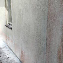 Michał Ostaszkiewicz - Elewacja budynku w trakcie mycia, widoczne spływające zanieczyszczenia i piana na kostce brukowej, fragment okna po lewej stronie.