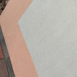 Michał Ostaszkiewicz - Fragment elewacji budynku z nałożonym tynkiem w kolorze łososiowym i szarym, widoczny fragment kostki brukowej.