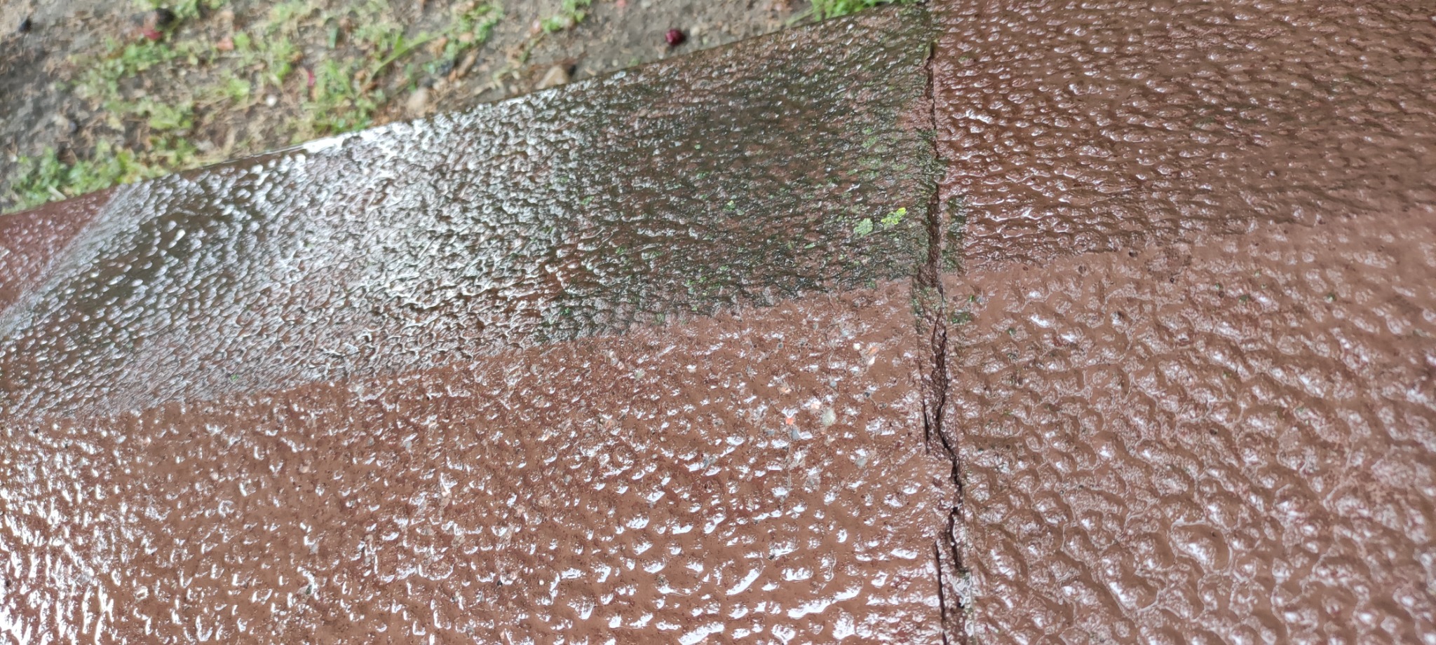 Częściowo umyta, brudna kostka brukowa w kolorze brązowym, z widocznym pęknięciem między kostkami i mchem w szczelinach.