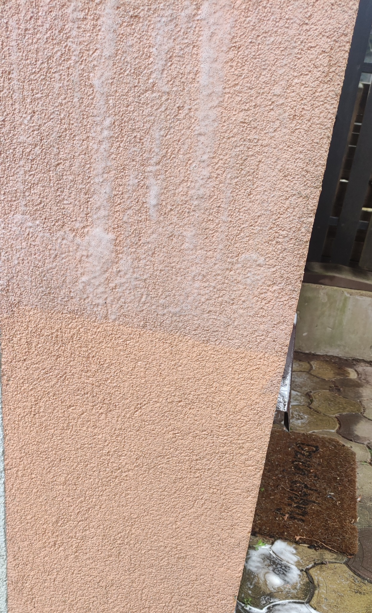 Fragment elewacji w kolorze brzoskwiniowym, z widoczną różnicą w czystości po myciu, zacieki z piany na kostce brukowej i wycieraczce.
