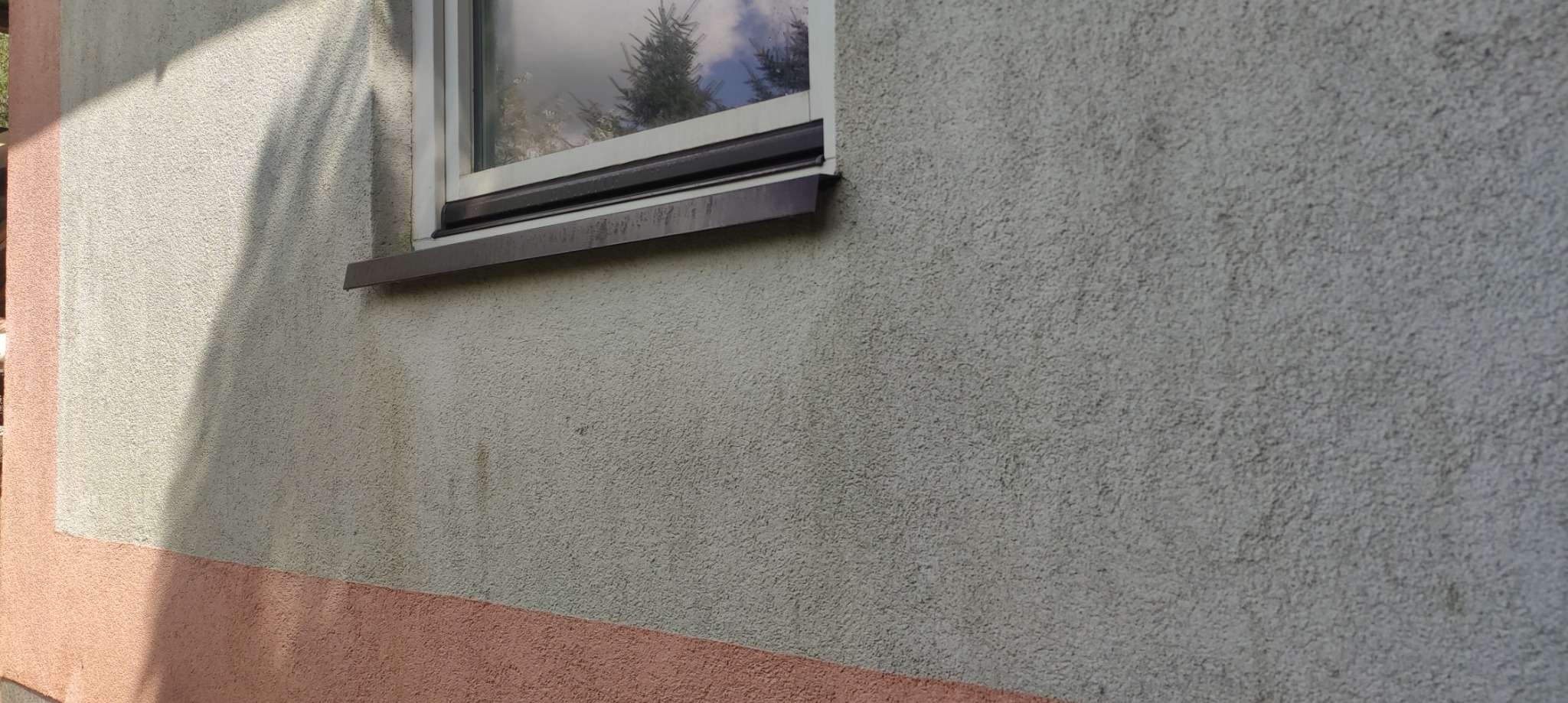 Szara, chropowata elewacja budynku z widocznym oknem i brązowym parapetem, z zaciekami i śladami zabrudzeń, dolna część ściany pomalowana na różowo.