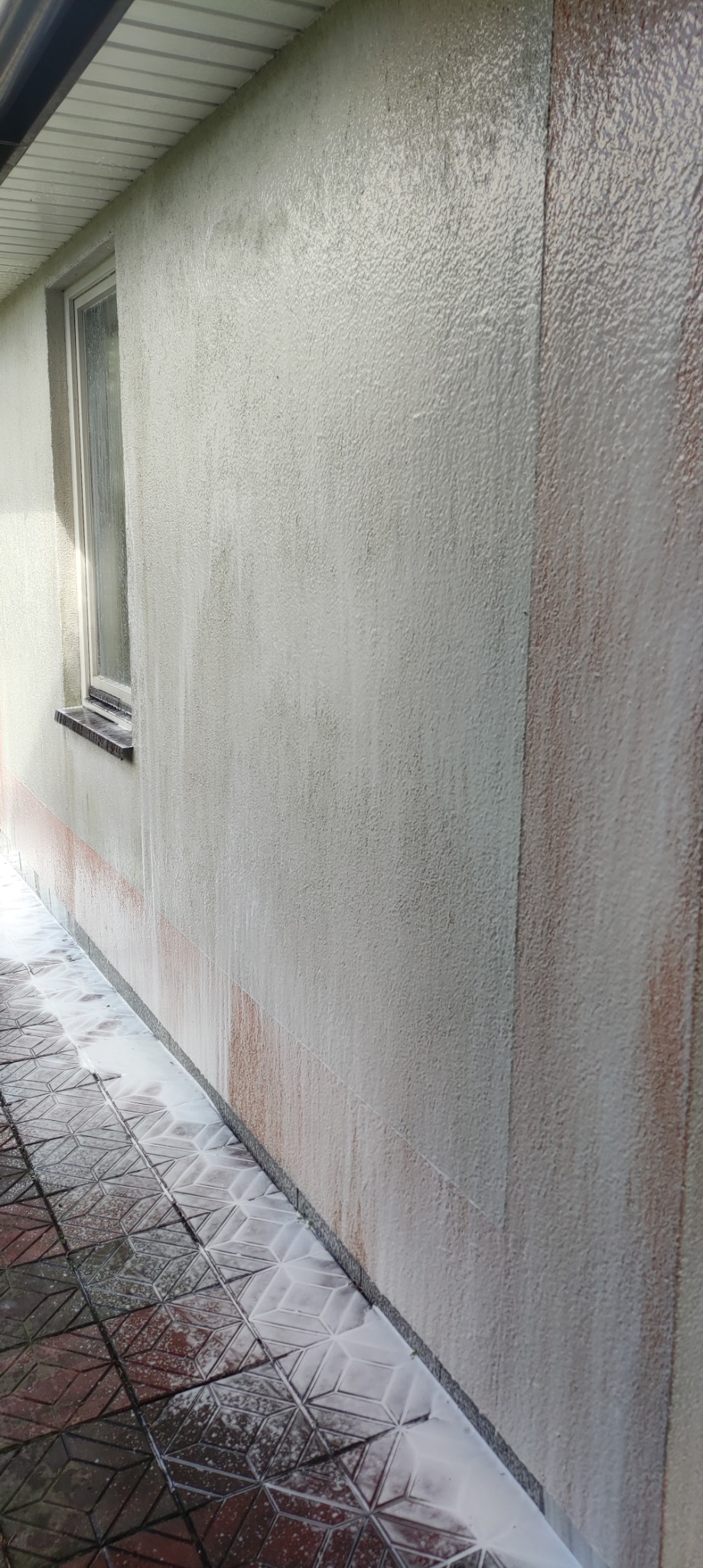 Elewacja budynku w trakcie mycia, widoczne spływające zanieczyszczenia i piana na kostce brukowej, fragment okna po lewej stronie.