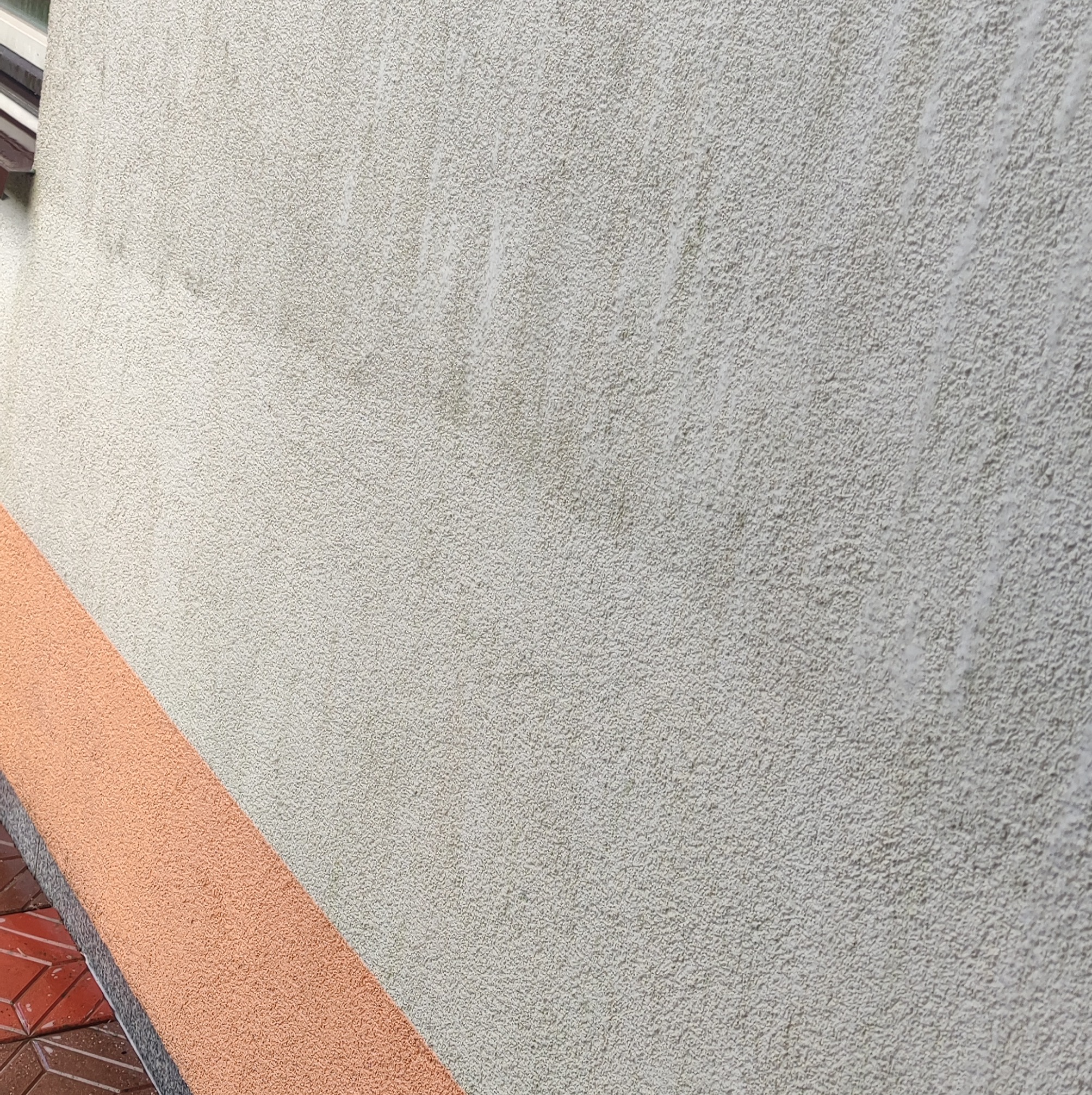 Szara elewacja z widocznymi śladami zabrudzeń i zacieków, z fragmentem pomarańczowego cokołu w dolnej części kadru.