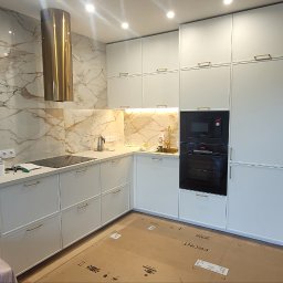 Rafał Kasprzyk - Biała kuchnia na wymiar z marmurowym backsplashiem, złotym okapem i sprzętem AGD w zabudowie, osłonięta kartonem i folią ochronną.