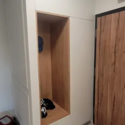 Biała szafa na wymiar z otwartą wnęką wykończoną drewnem, w której znajdują się kask rowerowy, czapka i buty; obok drzwi wejściowe z drewnopodobnym wzorem.