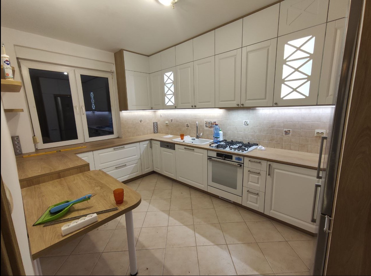 Jasna kuchnia w kształcie litery U z białymi frontami, drewnianymi blatami i podświetleniem LED pod szafkami wiszącymi; na stole leżą narzędzia i miarka.