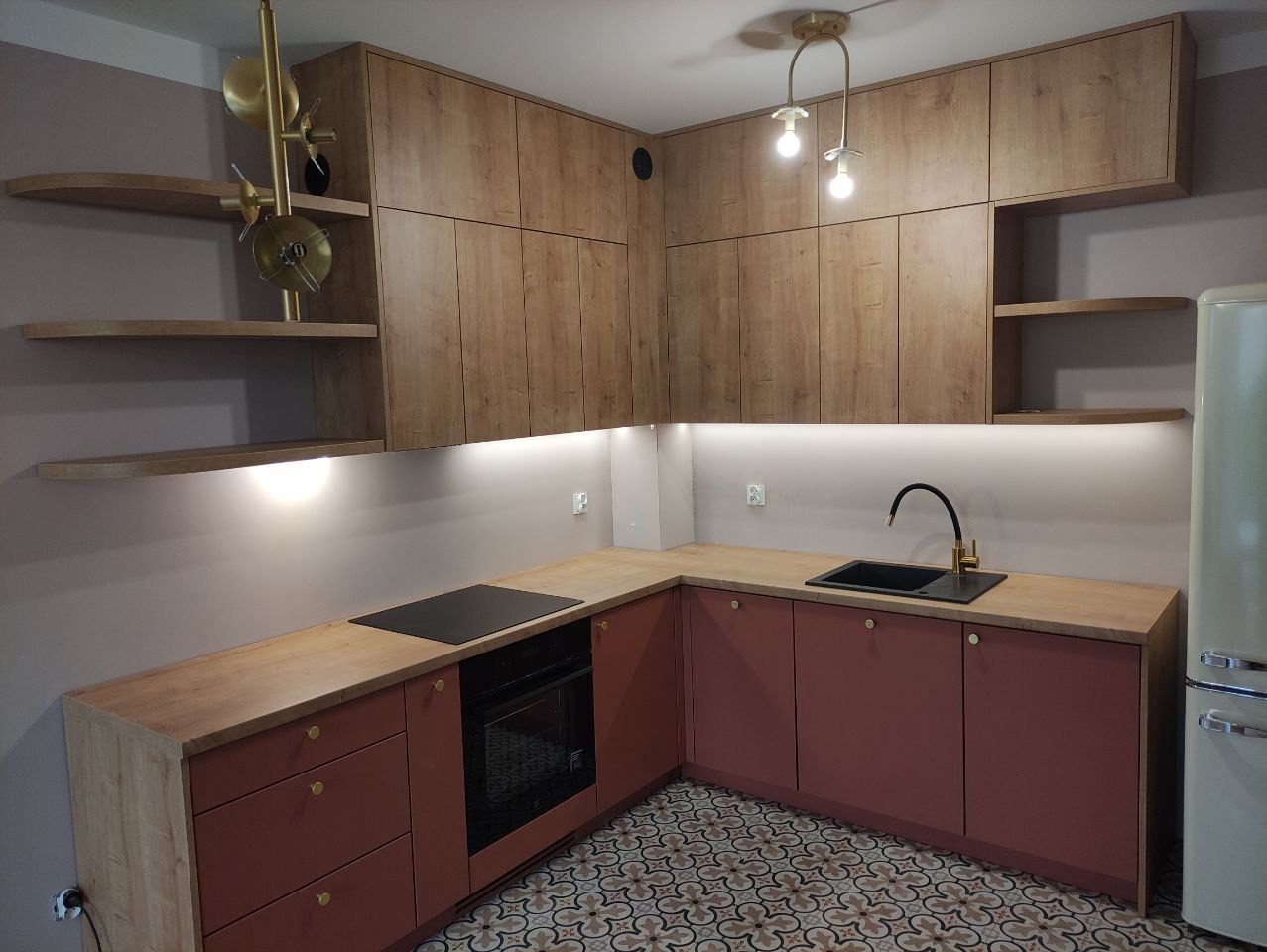 Nowoczesna kuchnia z drewnianymi szafkami górnymi i bordowymi dolnymi, blatem w kolorze jasnego drewna, czarnym zlewem i płytą indukcyjną, oświetleniem LED podszafkowym i dekoracyjną lampą sufitową.