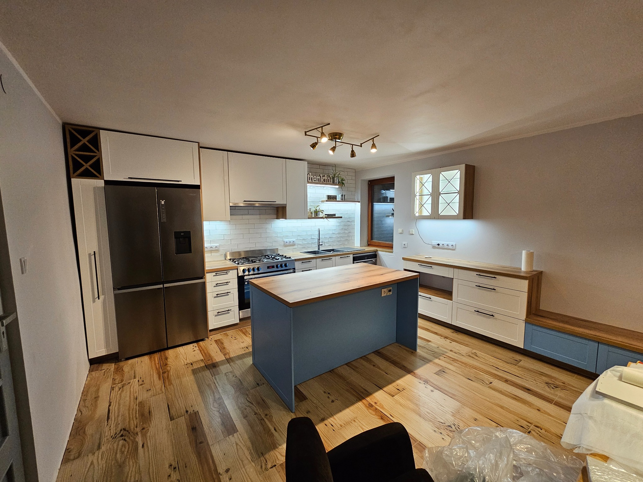 Jasna kuchnia z wyspą, drewnianą podłogą i białymi szafkami, oświetlona lampą sufitową z kilkoma punktami świetlnymi