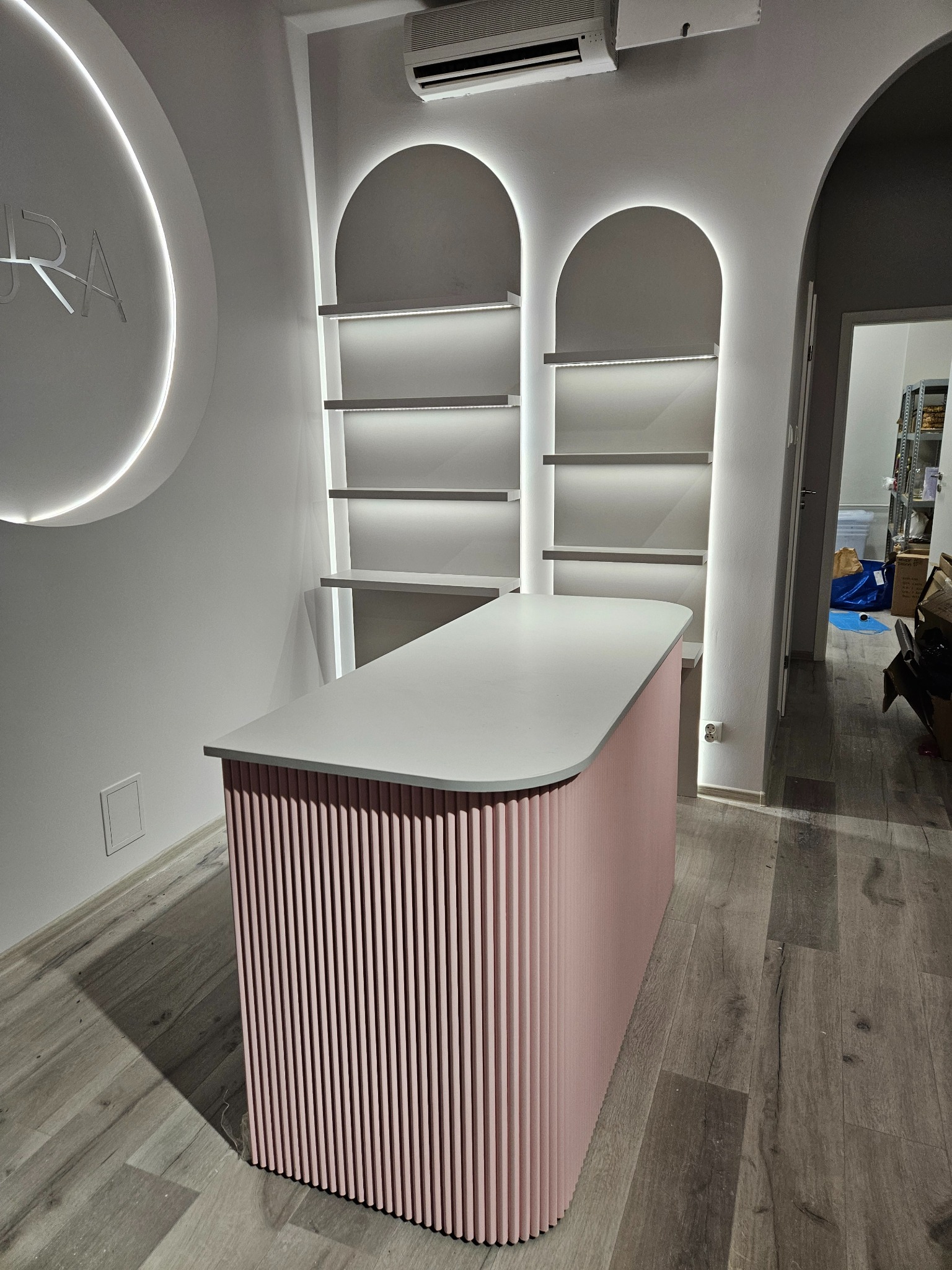 Nowoczesna lada recepcyjna z różowym, ryflowanym frontem i białym blatem, podświetlane półki w tle, okrągłe neonowe logo na ścianie.