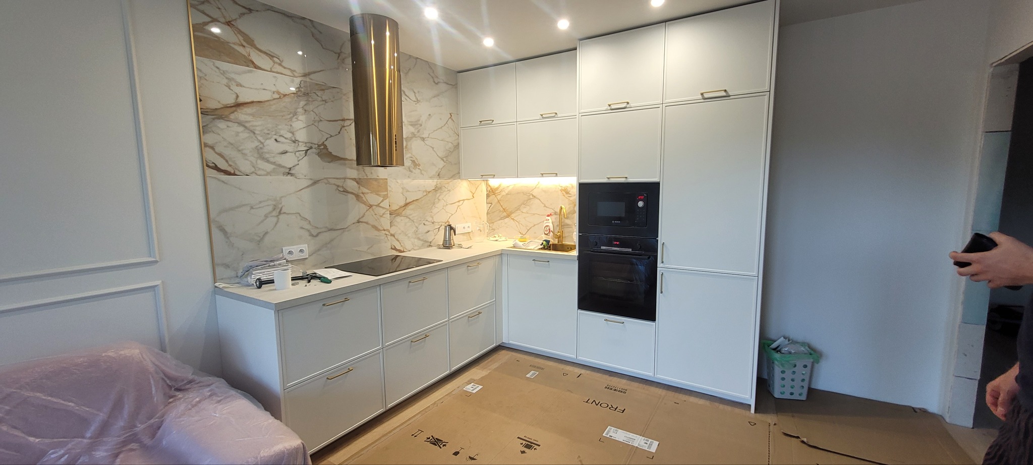 Biała kuchnia na wymiar z marmurowym backsplashiem, złotym okapem i sprzętem AGD w zabudowie, osłonięta kartonem i folią ochronną.