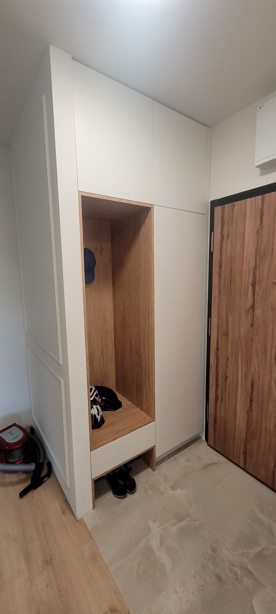 Biała szafa na wymiar z otwartą wnęką wykończoną drewnem, w której znajdują się kask rowerowy, czapka i buty; obok drzwi wejściowe z drewnopodobnym wzorem.