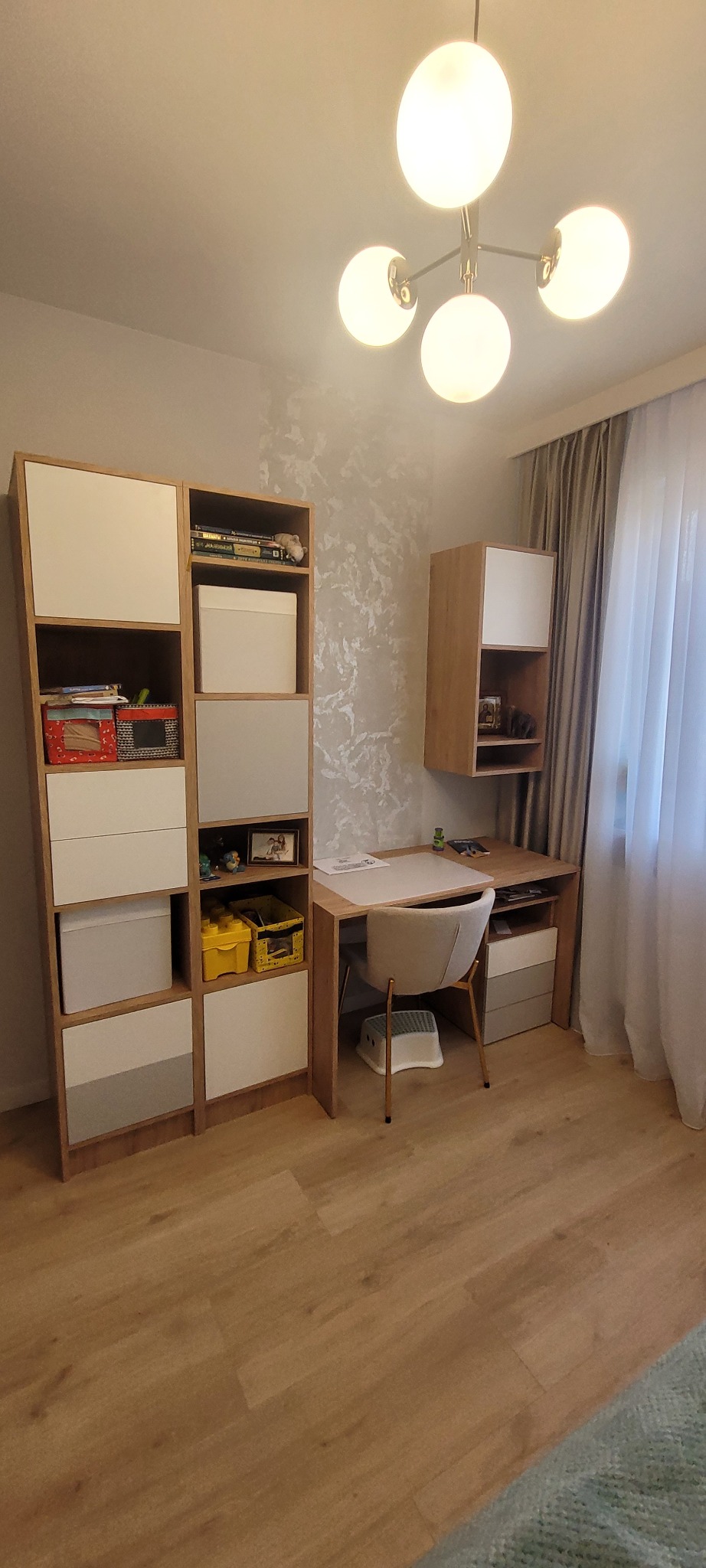 Pokój dziecięcy z biurkiem, krzesłem na złotych nóżkach i regałem z półkami oraz szafką wiszącą, oświetlony lampą sufitową z białymi kulami.