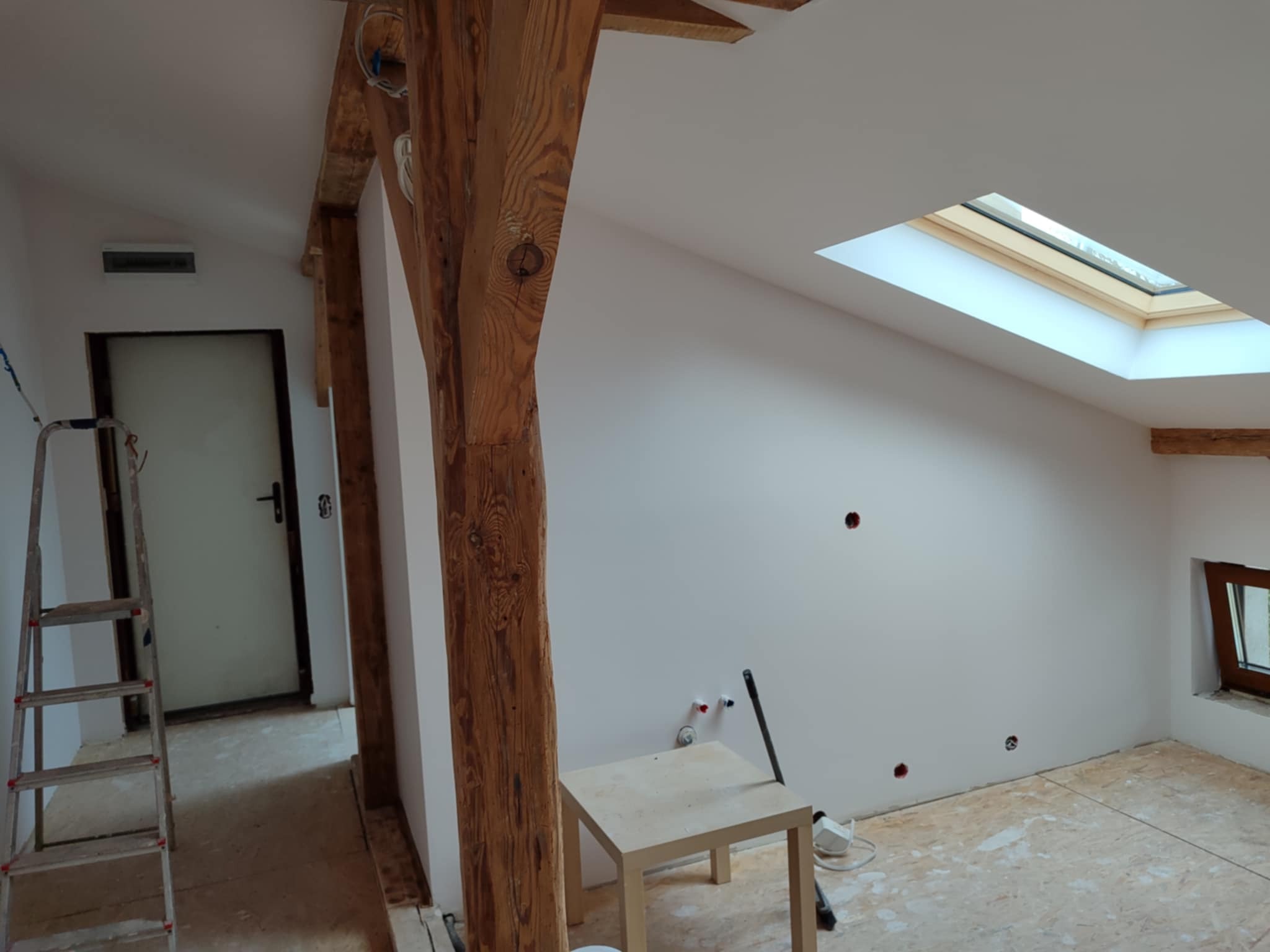 Poddasze po szpachlowaniu i malowaniu na biało, widoczne drewniane elementy konstrukcyjne, okno dachowe i drabina. Przygotowanie do dalszych prac wykończeniowych.