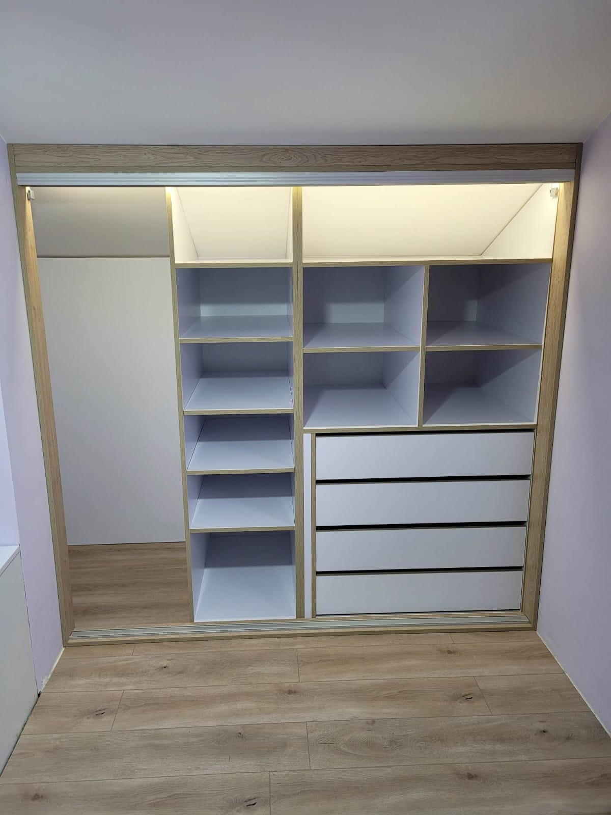 Wnętrze: szafa wnękowa z przesuwnymi drzwiami, oświetleniem LED i jasnym drewnianym obramowaniem, z widocznymi półkami i szufladami, na podłodze panele.