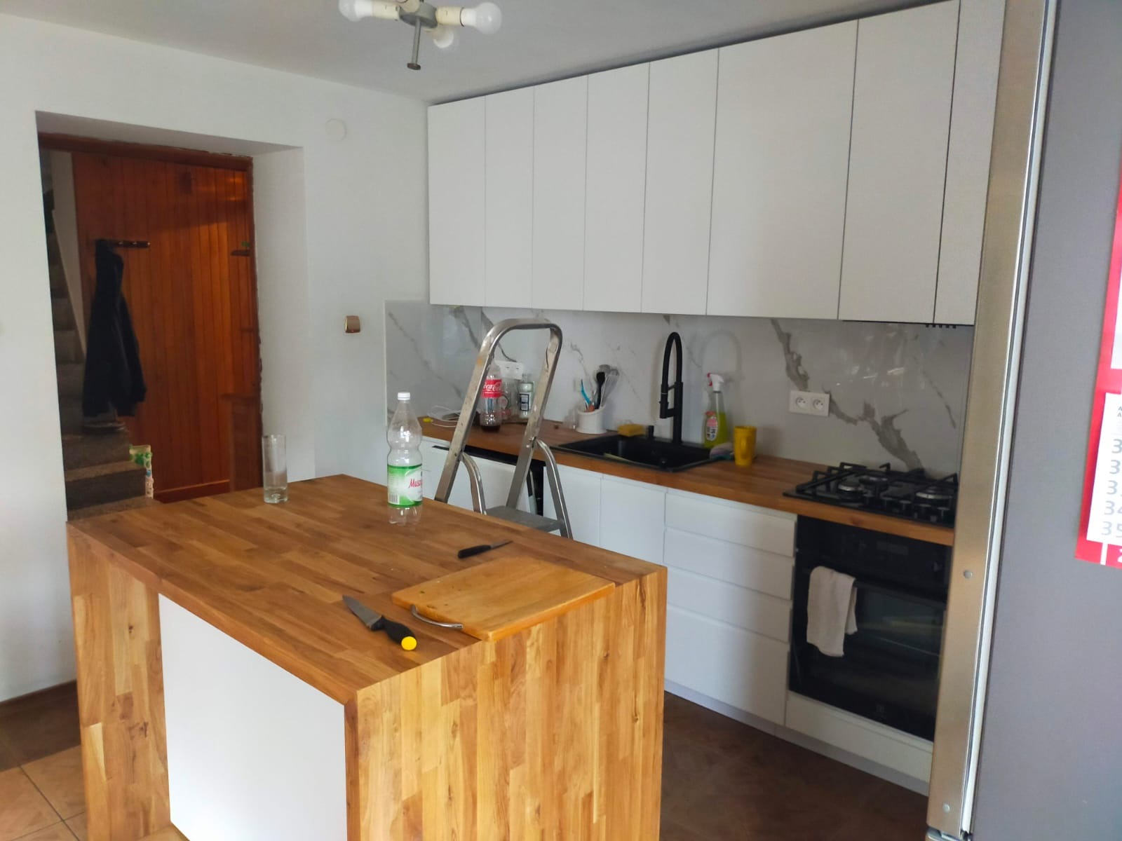 Kuchenne wnętrze z drewnianym blatem wyspy i białymi szafkami. Drabina oparta o ścianę. Widoczne akcesoria kuchenne i fragment wejścia do innego pomieszczenia.