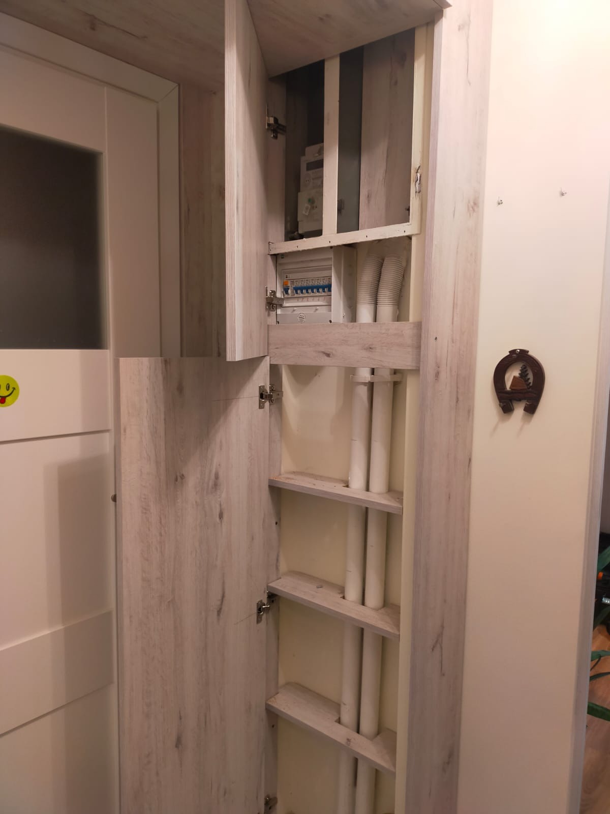 Otwarta szafka z półkami kryjąca instalację elektryczną i rury, wykończona w jasnym drewnie z widocznymi elementami mocowań.
