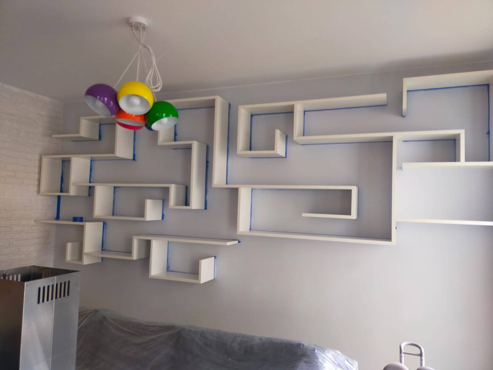 Białe, geometryczne półki na szarej ścianie, zabezpieczone niebieską taśmą malarską. Kolorowa lampa sufitowa. Dolna część kadru zasłonięta folią ochronną.