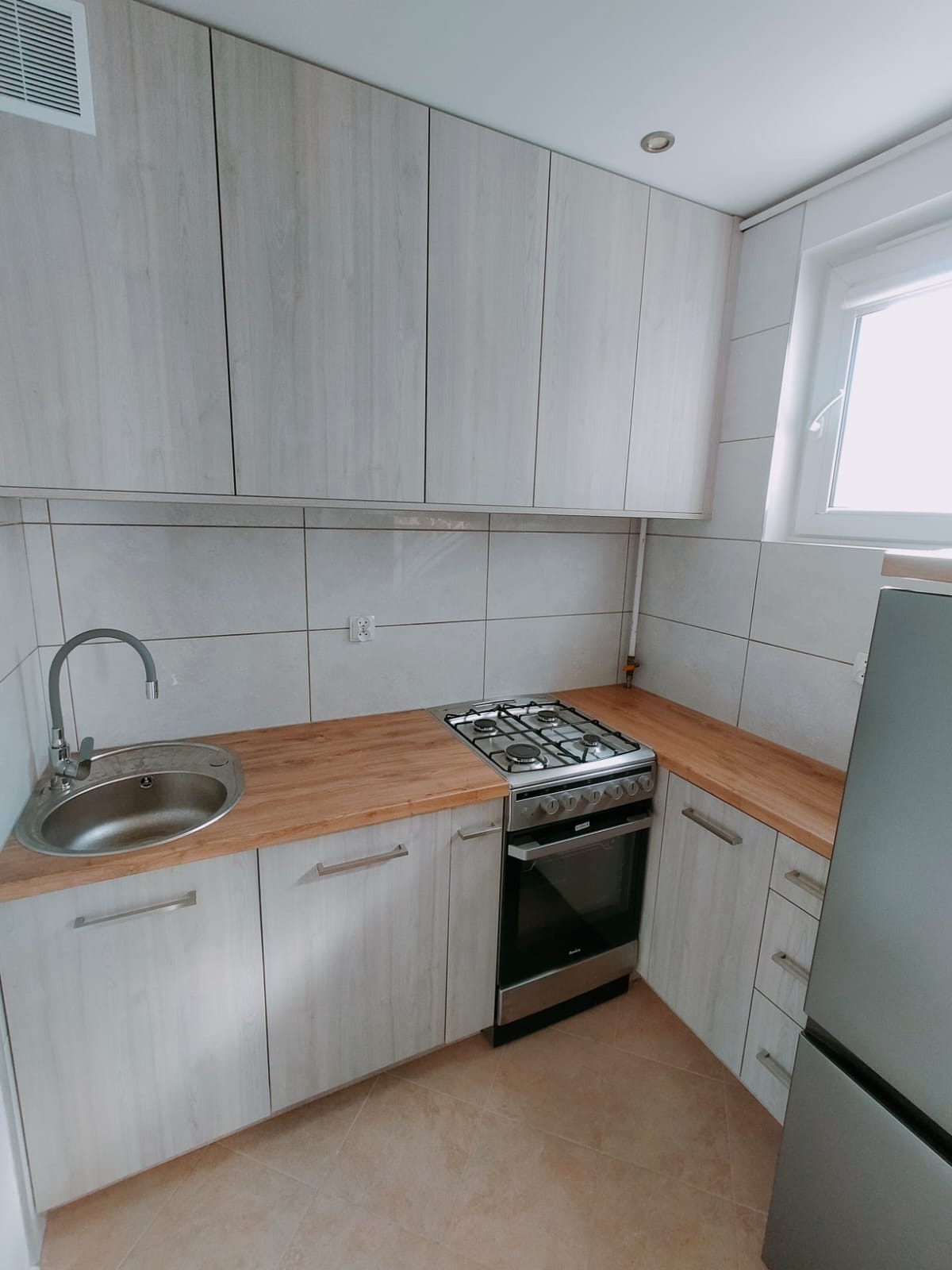 Jasna kuchnia z drewnianym blatem, srebrnym zlewem i kuchenką. Szafki w odcieniach bieli. Widoczne płytki na ścianie i fragment lodówki.