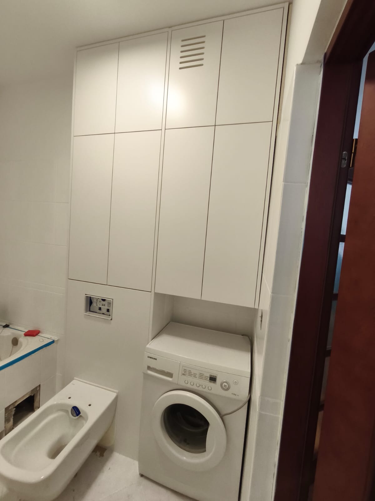 Biała zabudowa meblowa w łazience z pralką Samsung, bidetem i częściowo widoczną wanną. Minimalistyczny design, jasne wnętrze, płytki na ścianach.