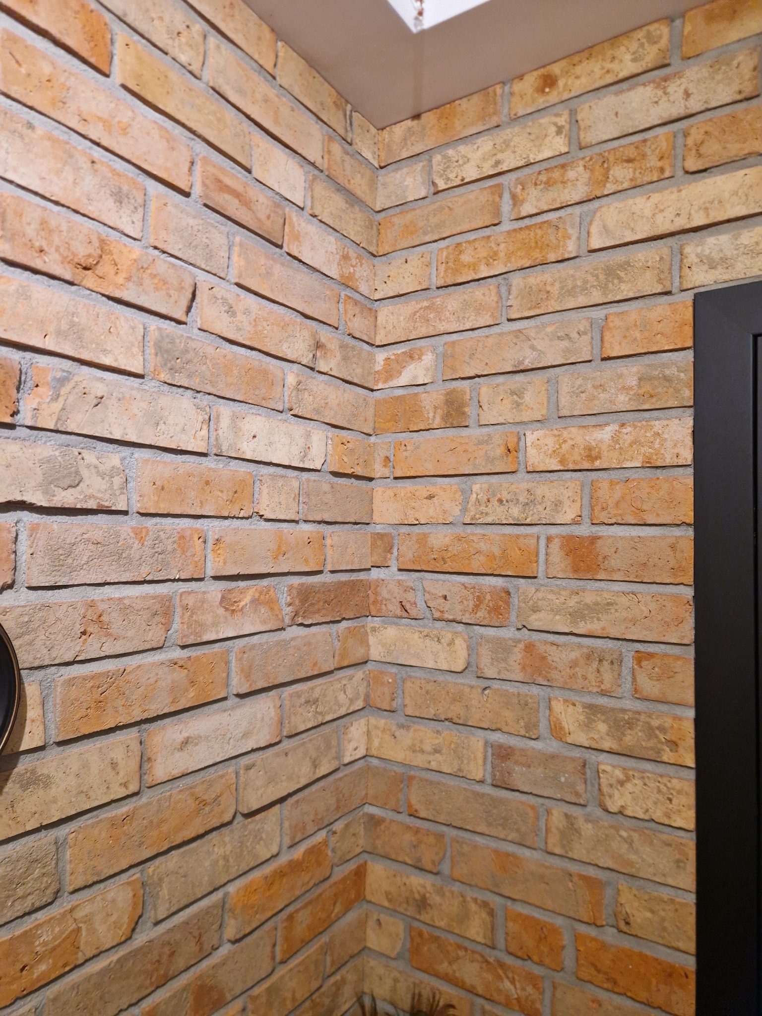 Dekoracyjna ściana wyłożona cegłą klinkierową w odcieniach beżu i pomarańczy, widoczny narożnik pomieszczenia i fragment czarnej ramy drzwi.