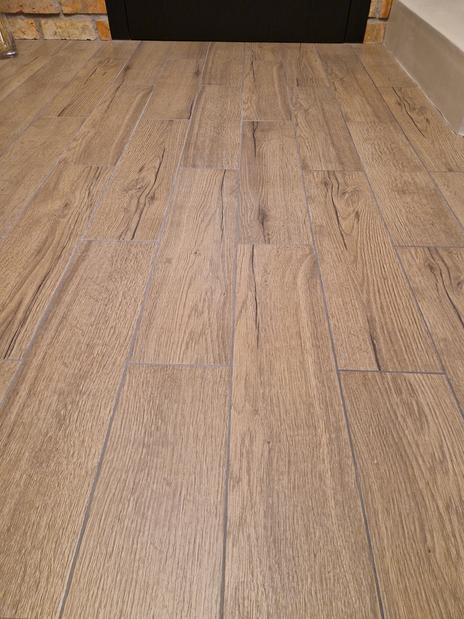 Ułożona podłoga z płytek imitujących drewno, widok z perspektywy podłogi, fuga w kolorze szarym, w tle fragment ściany z cegły.