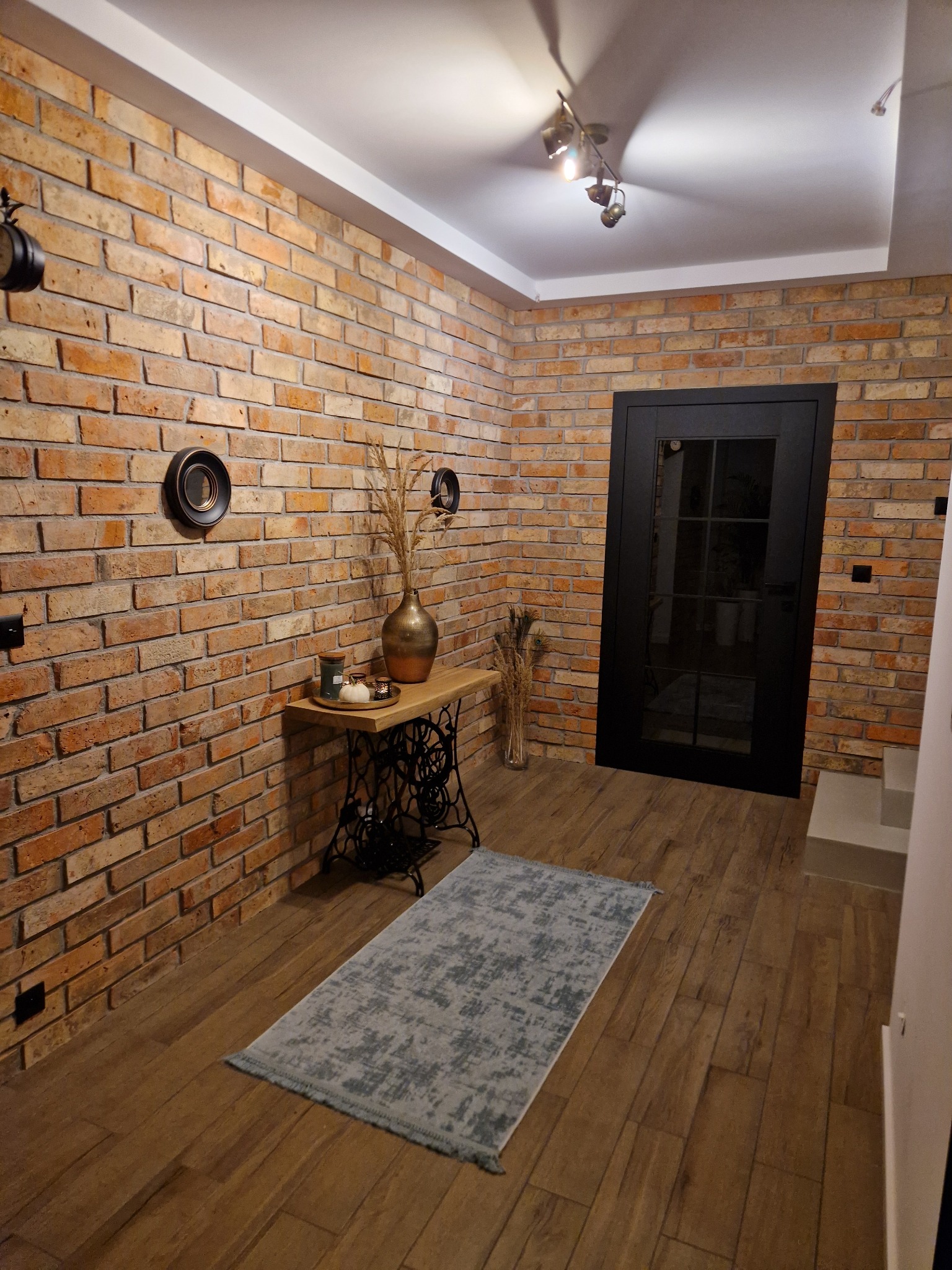 Wnętrze korytarza z ceglaną ścianą, drewnianą podłogą, czarnymi drzwiami i stolikiem ze starą maszyną do szycia, udekorowane wazonem z suszonymi trawami i okrągłymi dekoracjami ściennymi.